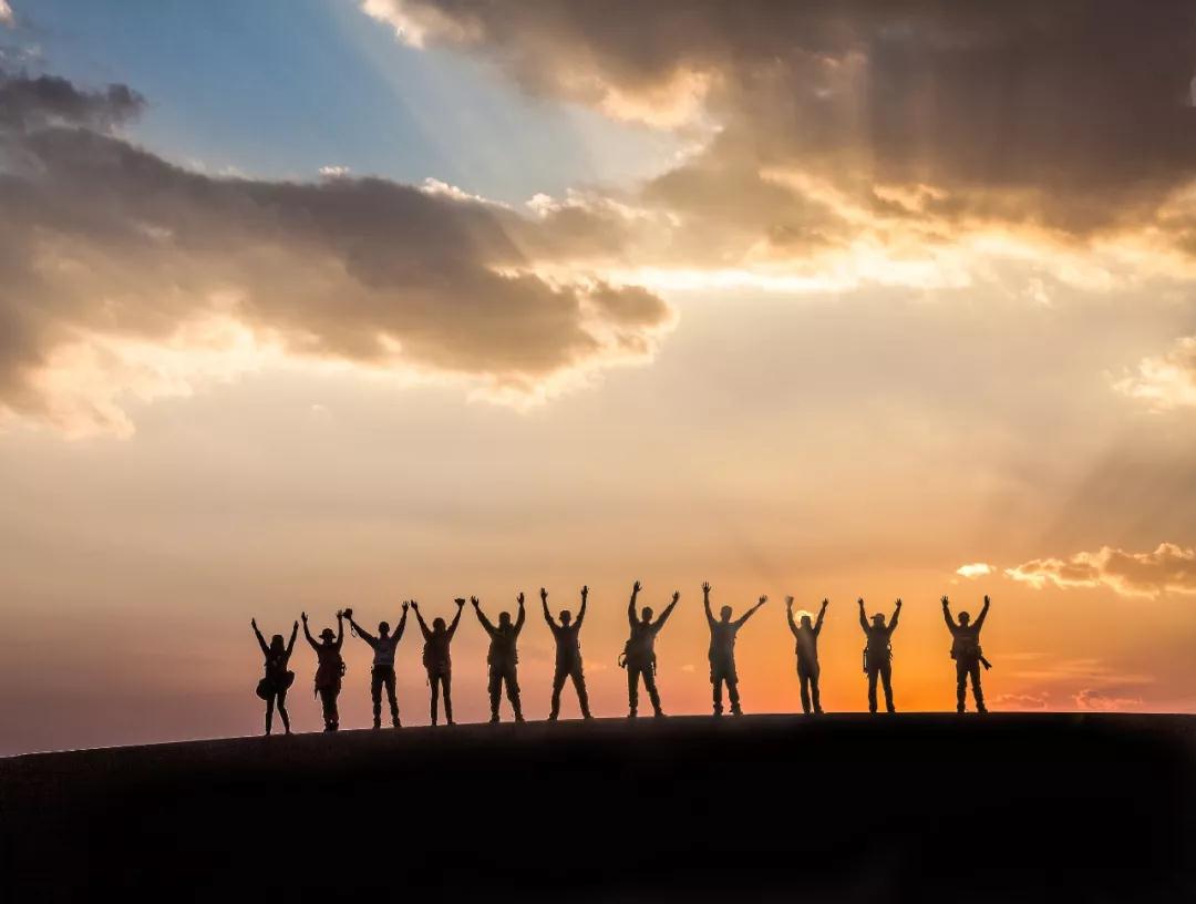 Silhouettes of a line of people with raised arms standing on a ridge against a colorful sunset sky