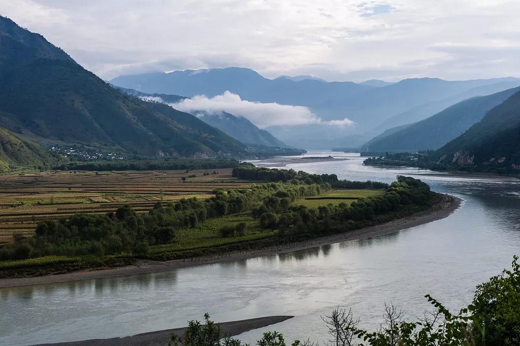 Wide river winding through a misty mountain valley with green fields and low clouds, calm blue-green tones